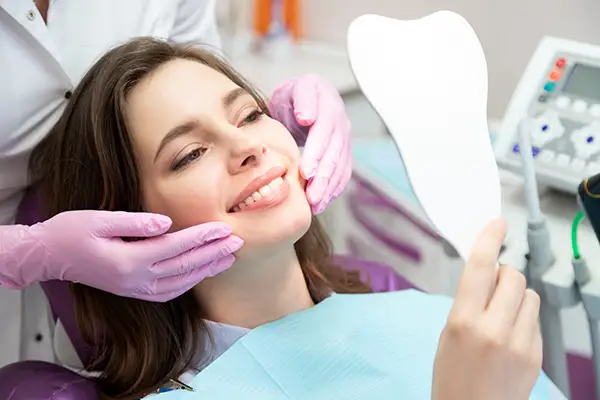 A smiling woman holding a tooth-shaped mirror while a dentist adjusts her face, showcasing her dental results in a clinic. A smiling woman holding a tooth-shaped mirror while a dentist adjusts her face, showcasing her dental results in a clinic.