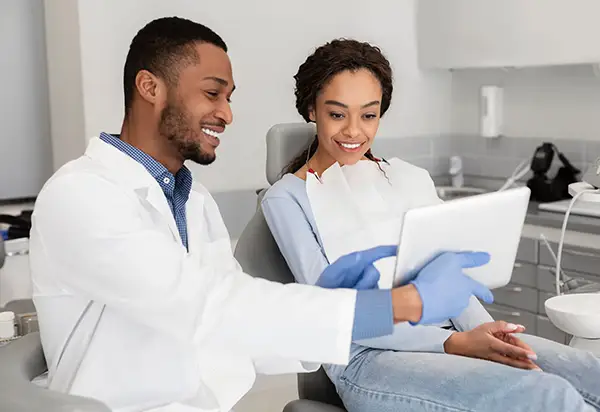 A smiling dental assistant presents a tablet with patient information to a content patient seated in a dental chair.